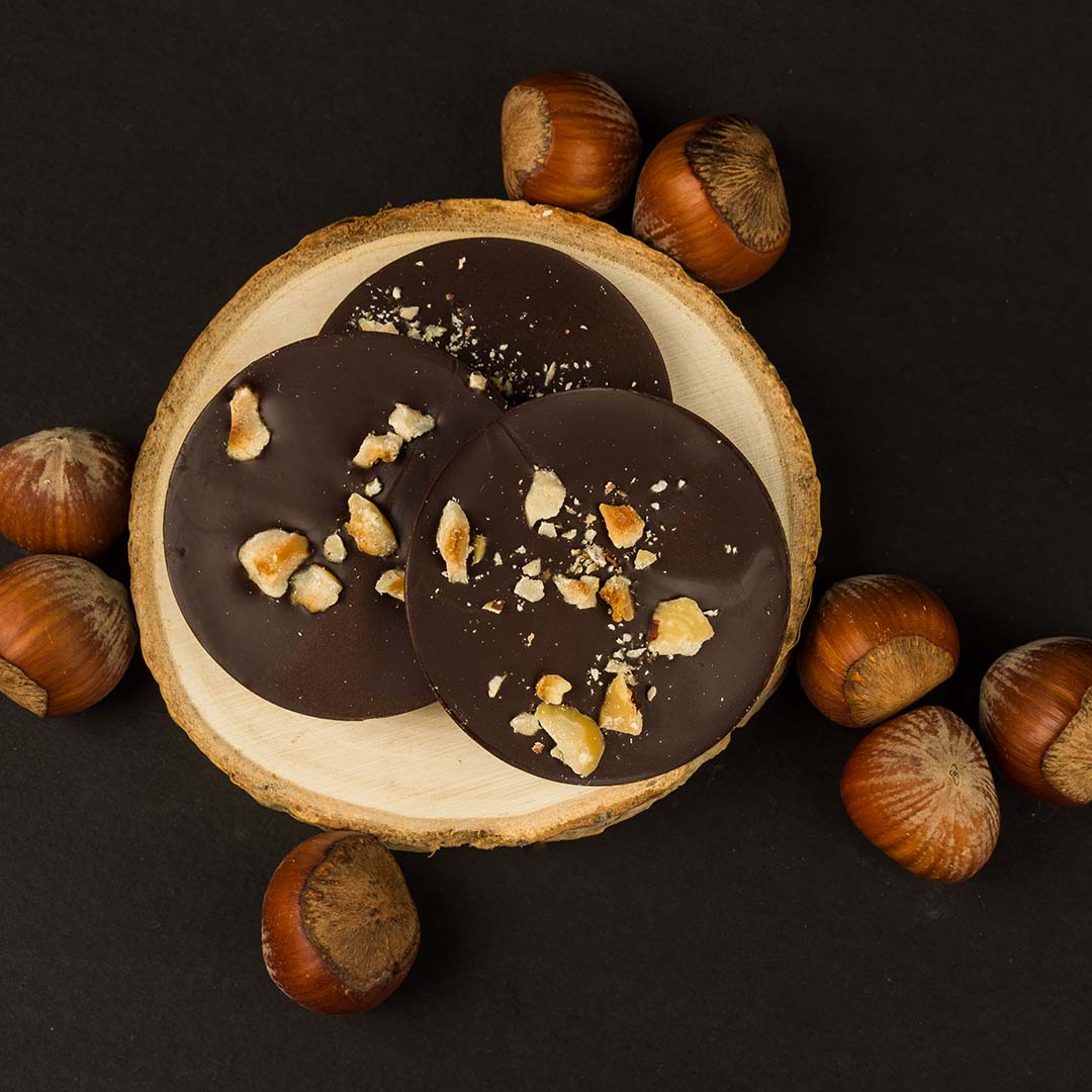 Three Allulose Chocolate discs with roasted hazelnut pieces displayed on a wood stage with whole hazelnuts all around them