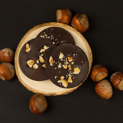 Three Allulose Chocolate discs with roasted hazelnut pieces displayed on a wood stage with whole hazelnuts all around them