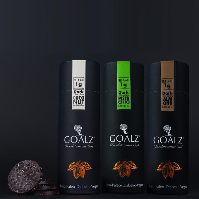 Three Goalz keto chocolate packs. First box is sugar free chocolate with coconut, second box is sugar-free dark chocolate with pistachio, third one is sugar-free chocolate with roasted Almonds. All shows their net carb as 1g. All three boxes show a text at the bottom saying Keto, Paleo, Diabetic, Vegan. On the left side there  are 4 chocolate discs stacked with some hazelnut flakes on them.
