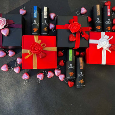 Black and Red chocolate gift boxes decorated with roses and hearts. Keto chocolate boxes with various flavors are also shown. The gift bozes are surrounded by heart shaped decorations on dark background 
